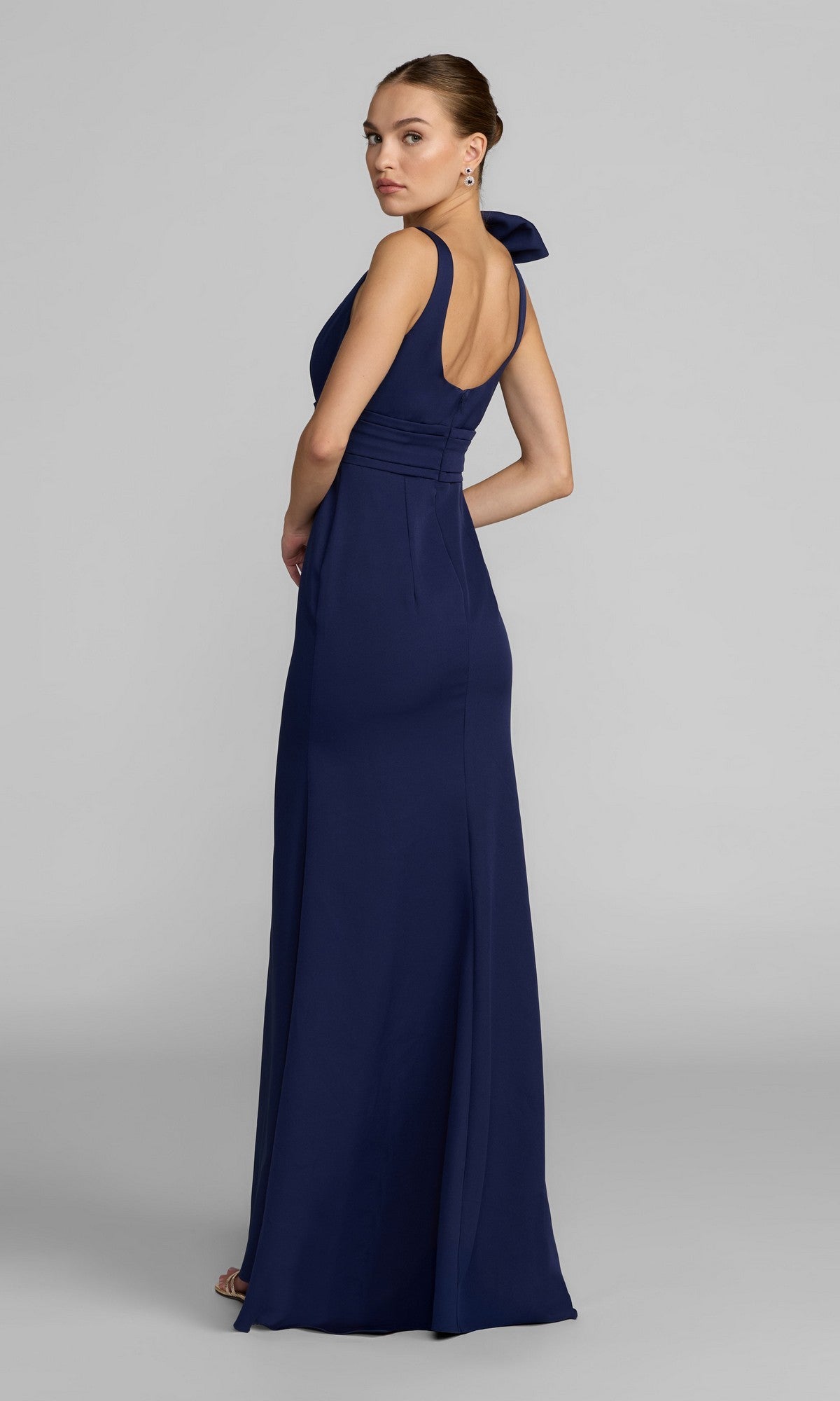 A woman poses sideways, glancing over her shoulder in the Mac Duggal 49964 Long Formal Dress—a floor-length, sleeveless navy crepe column gown with a fitted bodice—set against a plain gray background.